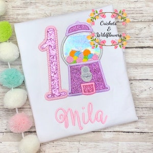 May include: A white onesie with a pink glittery number one and a pink glittery gumball machine with colorful gumballs. The name "Mila" is embroidered in pink below the gumball machine.