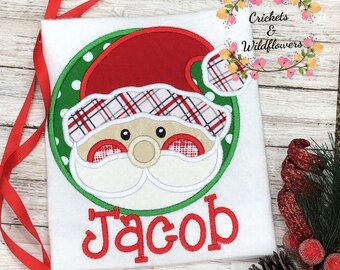 May include: A white t-shirt with a green circle containing a red and white plaid Santa Claus face. The name "Jacob" is embroidered in red below the circle.