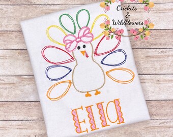 May include: A white t-shirt with an embroidered turkey design. The turkey has a pink bow and colorful feathers. The name "Ellie" is embroidered below the turkey.