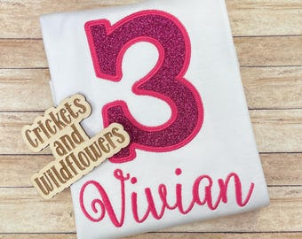 May include: A white t-shirt with a pink glittery number 3 and the name Vivian embroidered in pink. A wooden sign with the text "Crickets and Wildflowers" is in the lower left corner.