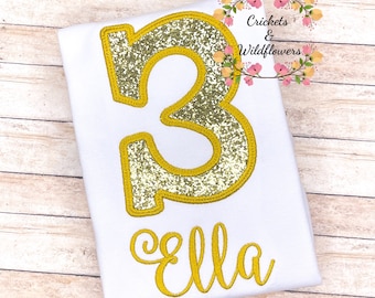 May include: A white t-shirt with a gold glitter number 3 and the name "Ella" embroidered in gold thread. The shirt is for a child's birthday.