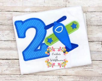 May include: A white t-shirt with a blue number 2 and a blue airplane with green wings and white stars. The text "Crickets & Wildflowers" is below the airplane.