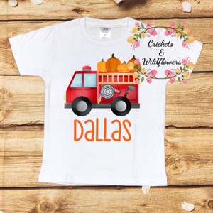 May include: A white short-sleeve t-shirt featuring a graphic of a red fire truck carrying orange pumpkins. The name DALLAS is printed in orange letters below the truck. A floral wreath logo with the text Crickets & Wildflowers is visible.