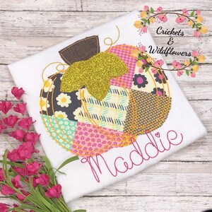 May include: A white t-shirt with a patchwork pumpkin design embroidered on the front. The pumpkin is decorated with a gold glitter stem and the name "Maddie" is embroidered below it. The shirt is surrounded by pink flowers and a wooden background.