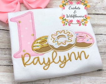 May include: A white onesie with a pink number one, three embroidered donuts, and the name "Raylynn" in gold thread. The onesie is decorated with pink polka dots.
