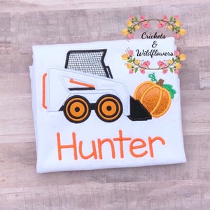 May include: White and orange embroidered design of a small construction vehicle with a pumpkin on a white t-shirt. The name "Hunter" is embroidered in orange below the design.