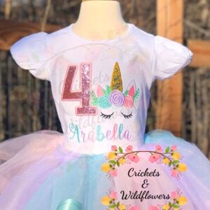 以下が含まれることがあります： ピンクのキラキラした数字の4と、花が刺繍されたユニコーンが描かれた白いTシャツ。Tシャツにはユニコーンの下に「Arabella」と書かれています。Tシャツはレインボーのチュチュスカートと組み合わせています。