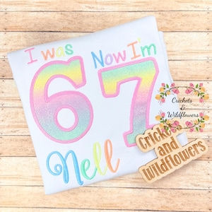 May include: White t-shirt with the embroidered text "I was 67 Now I'm" in pastel shades. The name "Nell" is embroidered below the number 67. A wooden sign with the text "Crickets and Wildflowers" is on the right.