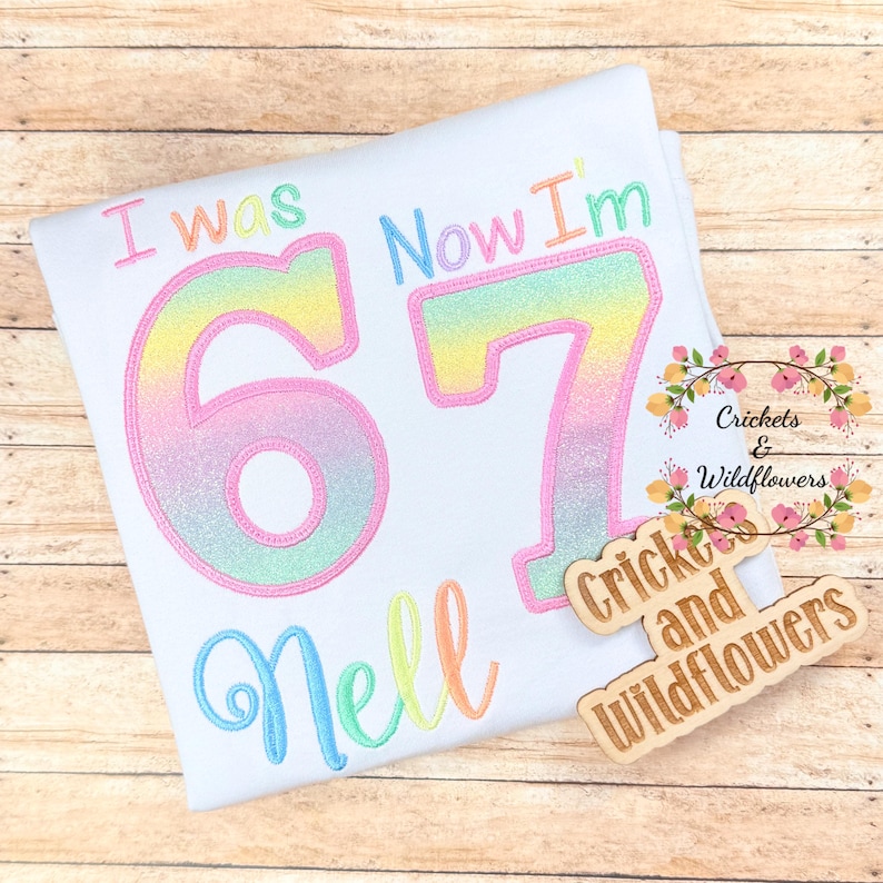May include: White t-shirt with the embroidered text "I was 67 Now I'm" in pastel shades. The name "Nell" is embroidered below the number 67. A wooden sign with the text "Crickets and Wildflowers" is on the right.