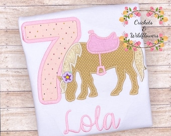 May include: White t-shirt with a pink and gold embroidered design. Features a large number 7, a pony with a saddle, and the name "Lola" in pink script. The pony is tan with a gold mane and tail. The text "Crickets & Wildflowers" is in the upper right corner.