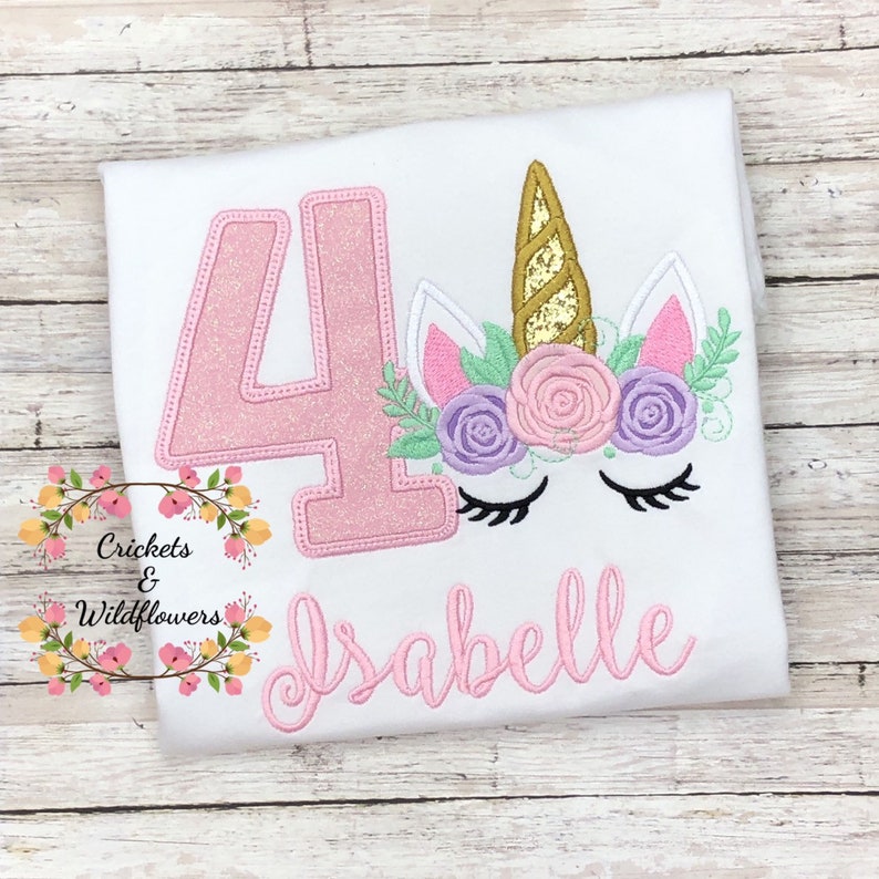 May include: White t-shirt with a pink glittery number 4 and a unicorn face design. The unicorn has a gold horn, pink and purple flowers, and the name "Isabelle" in pink script. The shirt has the text "Crickets & Wildflowers".