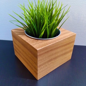 May include: A square wooden planter box with a light brown wood grain finish. The planter contains a green artificial plant with long, thin leaves.