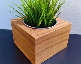 May include: A wooden planter box with a square shape and a light brown wood grain finish. The planter contains a green artificial plant with long, thin leaves.