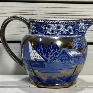 Puede incluir: Una jarra de cerámica azul y blanca con acabado plateado. La jarra presenta una escena de cabaña con árboles y un techo nevado.