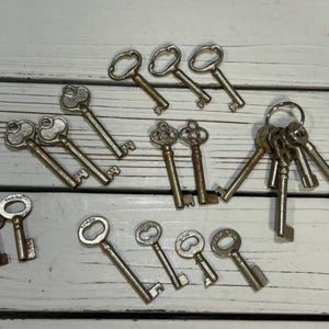 May include: A collection of antique keys in various shapes and sizes, arranged on a white wooden surface. The keys are made of metal and show signs of age, with some having ornate designs and others with simple, functional forms. A few keys are attached to a metal ring.