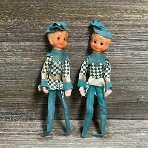 Op de afbeelding: Twee elfpoppen met beschilderde gezichten, blauwe ogen en roze wangen. Elke pop draagt een blauwgroene puntmuts, een blauwgroene kraag en een tuniek met lange mouwen met een wit en blauwgroen ruitpatroon en een blauwgroene broek.