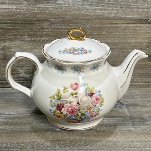 May include: Cream-colored porcelain teapot with a floral design featuring pink roses and white doves. The lid has a gold handle and is decorated with small blue and pink flowers. Gold trim accents the handle and spout.