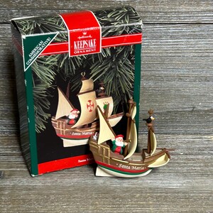 May include: A Hallmark Keepsake ornament depicting the Santa Maria ship with Santa Claus figures. The ornament is brown, beige, and red, with the words "Santa Maria" on the side. The box is green and red, with the words "American Commemorative" and "Keepsake Ornament".