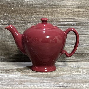 Può includere: Una teiera in ceramica bordeaux con coperchio e manico. La teiera ha un corpo arrotondato, un beccuccio curvo e un manico ad anello. La teiera è su una superficie di legno.