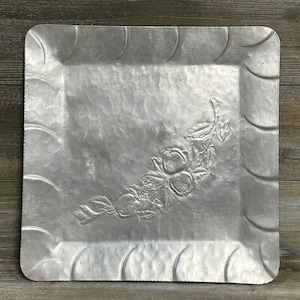 May include: A square, silver-toned metal tray with a floral design. The tray has a raised, scalloped edge and a detailed embossed pattern of flowers and leaves in the center. The tray is likely used for serving or display.