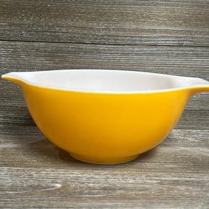 May include: A vintage, bright yellow mixing bowl with a white interior. The bowl has a rounded shape with a flared rim and two small handles. The bowl is made of ceramic or glass and is likely from the mid-20th century.