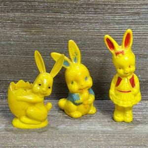 Może przedstawiać: Trzy żółte figurki królików z tworzywa sztucznego. Lewy królik siedzi w żółtej skorupce jajka. Środkowy królik ma niebieskie akcenty na uszach i klatce piersiowej. Prawy królik stoi prosto, ubrany w czerwoną kamizelkę i czerwoną spódnicę.