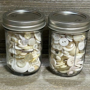 May include: Two glass jars filled with a variety of off-white and cream-colored buttons. The buttons are different sizes and shapes, with two- and four-hole designs. The jars have silver lids and are displayed on a wooden surface.