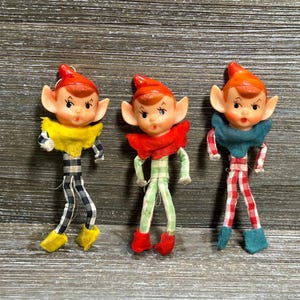 May include: Three elf figurines with pointed orange hats and painted facial features. Each elf wears a fabric collar and matching patterned pants. One elf has a yellow collar and black-and-white plaid pants, one has a red collar and green-and-white plaid pants, and one has a teal collar and red-and-white plaid pants.