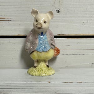 May include: A ceramic figurine of a pig dressed in a light purple jacket, blue shirt, and yellow pants, holding a small orange basket. The pig stands on a green base with grass details.
