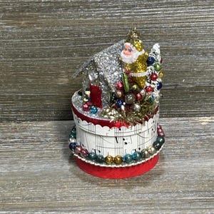 May include: A round decorative box wrapped in sheet music paper with a red base and a border of multicolored beads. Atop the box sits a silver glittered house, a small Santa Claus figure in a gold suit, and a cluster of colorful miniature ornaments.