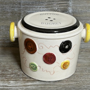 Op de afbeelding: Een crèmekleurige keramische knopemmer met een deksel, zwarte rand en gele handvatten. De emmer is versierd met verschillende gekleurde knopontwerpen en de woorden "BUTTON BUCKET" op het deksel.