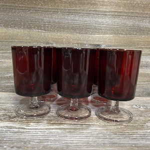 May include: Six ruby red stemmed glasses. The glasses have a cylindrical bowl and a clear, circular base. The stem is clear and connects the bowl to the base. The glasses are arranged on a wooden surface.