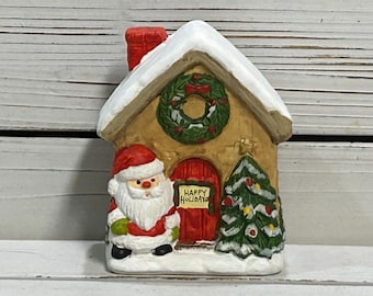 May include: A ceramic Christmas house figurine with a snow-covered roof, a red door, and a green wreath. Santa Claus stands beside the house, and a Christmas tree is on the right. The sign on the door reads "Happy Holidays."