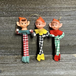 May include: Three elf ornaments with pointed orange hats and painted faces. Each elf wears a felt collar and a long, thin body covered in gingham fabric. The first elf has a red and white pattern, the second black and white, and the third green and white.