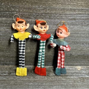 May include: Three vintage-style elf ornaments with pointed ears and painted faces. Each elf wears a felt hat and a matching collar, with long, thin limbs covered in chequered fabric in black and white, green and white, or red and white patterns.