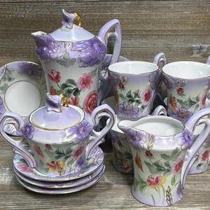 Peut inclure: Service à thé en porcelaine vintage dans les tons lavande, blanc et or. L'ensemble comprend une théière, un sucrier, une crémière, des tasses et des soucoupes, tous décorés de motifs floraux. Un charmant ajout à toute fête de thé.