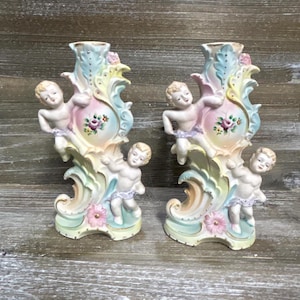 Może przedstawiać: Para świeczników porcelanowych z pastelowymi zdobieniami w stylu rokoko i motywami kwiatowymi. Każdy świecznik ozdobiony jest dwiema figurkami aniołków, umieszczonymi w górnej części oraz u podstawy, pomalowanymi w delikatne odcienie różu, błękitu i zieleni.