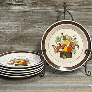 Puede incluir: Juego de platos de cerámica vintage con borde marrón y diseño de cesta de frutas. Los platos están apilados y expuestos en un soporte de metal, mostrando una estética retro. Los platos tienen aproximadamente 25 cm de diámetro.