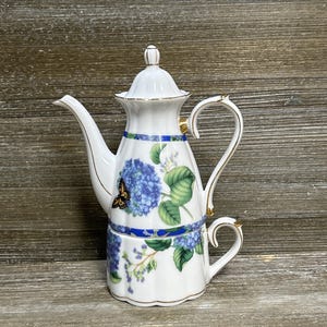 Może przedstawiać: Biały porcelanowy zestaw do herbaty dla jednej osoby, składający się z czajniczka ustawionego na filiżance. Zestaw ozdobiony jest niebieskimi hortensjami, zielonymi liśćmi i małym motylem. Krawędzie uchwytu, dzióbka i pokrywki są wykończone złotem.