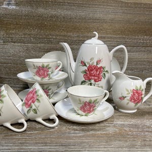 May include: A white porcelain tea set with a floral rose design. The set includes a teapot, creamer, sugar bowl, and several teacups and saucers. The roses are pink and red with green leaves, and the rims are trimmed in silver.