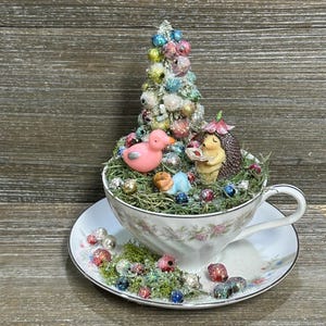 May include: A decorative teacup and saucer set filled with a miniature fairy garden. The garden includes a glittery tree, a pink duck, a hedgehog, and a sleeping baby figurine, all nestled in moss and colorful beads.
