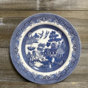Puede incluir: Un plato redondo de cerámica azul y blanco con un diseño detallado de patrón Willow. El plato presenta una escena central con pagodas, puentes y pájaros, rodeada por un borde decorativo. El plato está sobre un fondo de madera rústica.