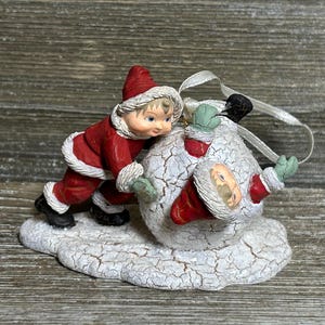 Könnte beinhalten: Weihnachtsschmuck mit einem Kind in rotem Anzug und Hut, das einen Schneemann schiebt. Der Schneemann trägt einen roten Anzug und Hut. Der Schmuck hat ein weißes Band zum Aufhängen und steht auf einer schneebedeckten Basis.