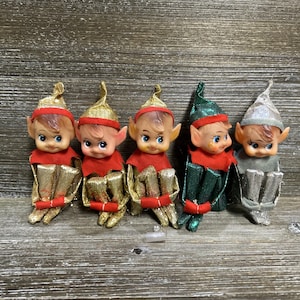 May include: Five small elf figurines with pointed ears and painted facial features. Three elves wear gold hats and outfits, one wears a green hat and outfit, and one wears a silver hat and outfit. Each elf has a red collar and red trim on their clothing.