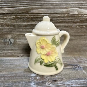 May include: Cream-colored ceramic teapot with a lid and handle, adorned with yellow and pink floral designs and green leaves. The teapot has a spout for pouring and is likely used for serving tea or other beverages.