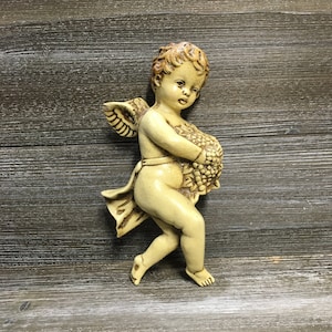 Puede incluir: Una figurita de querubín vintage con alas, sosteniendo un ramo de flores. La figurita está hecha de un material beige claro y tiene un aspecto rústico y antiguo.