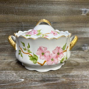 May include: A white ceramic serving dish with a lid, decorated with pink floral patterns and gold accents. The dish has a scalloped edge and gold handles. The lid features a gold handle and matching floral pattern. A decorative serving piece.