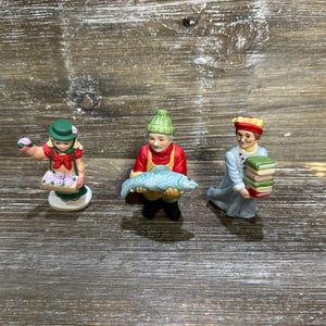 May include: Three ceramic figurines standing in a row. The left figure wears a green hat and holds a basket of purple flowers. The center figure wears a green knit cap and holds a large blue fish. The right figure wears a red hat and holds a stack of books.