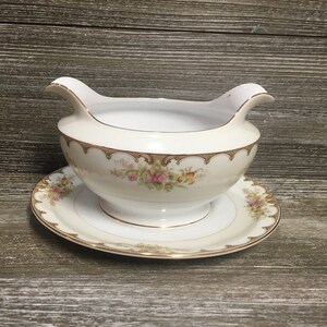 Puede incluir: Una salsera de porcelana blanca con ribete dorado y estampado floral. La salsera está sobre un plato a juego.