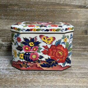 Può includere: Una scatola di metallo ottagonale con sfondo color crema e un vivace motivo floreale. Il design include grandi peonie rosse, fiori più piccoli rosa e gialli, foglie blu scuro e una farfalla gialla con dettagli rosa e neri.